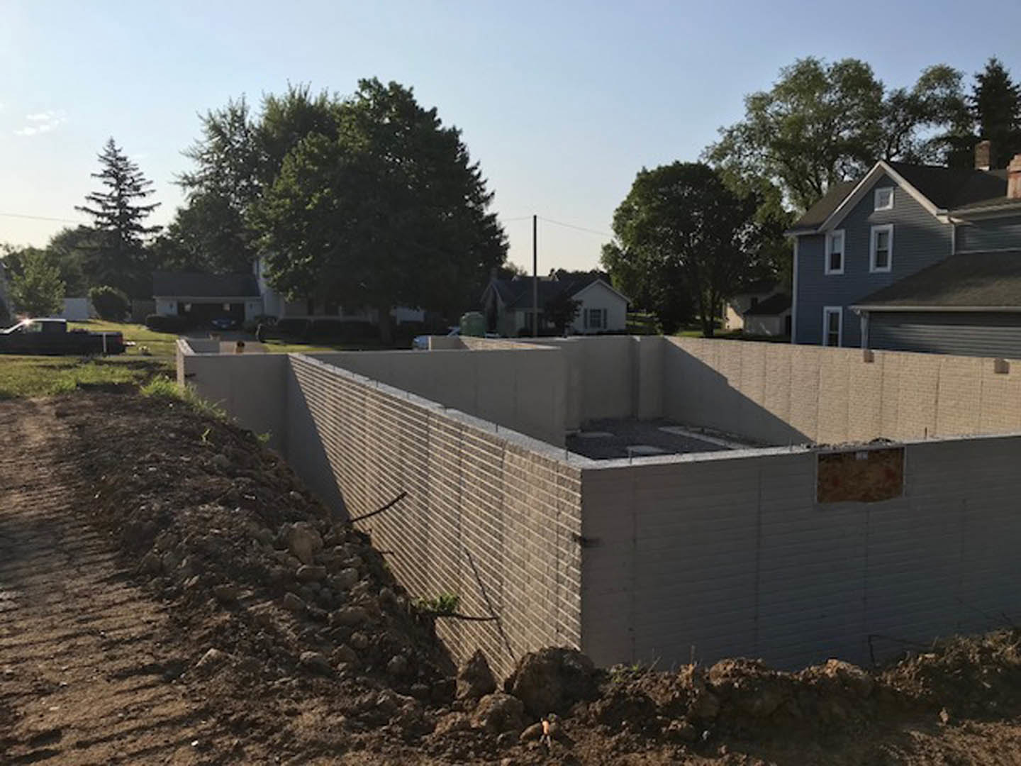 Framed custom home under construction with exposed composite walls, dirt mound in foreground, and mature trees surrounding property