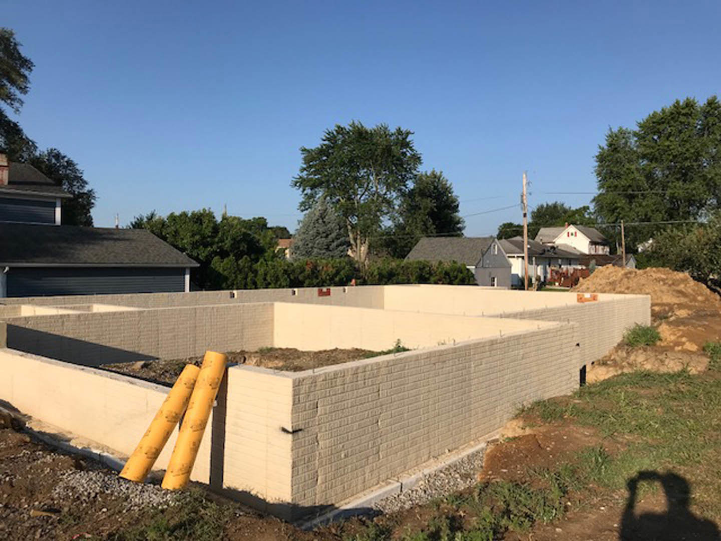 Concrete house foundation with exposed yellow pipes, brick wall, patch of dirt, leafy tree, and blue sky in the background