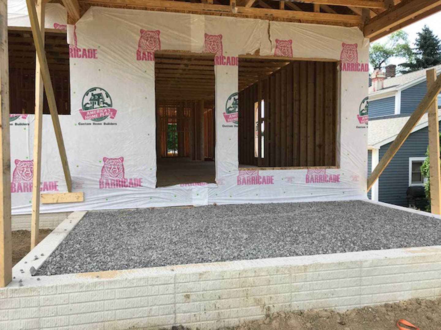Unfinished house exterior with white plastic sheeting covering walls, exposed wooden beams, gravel driveway, and visible pink and grey construction materials near a window.
