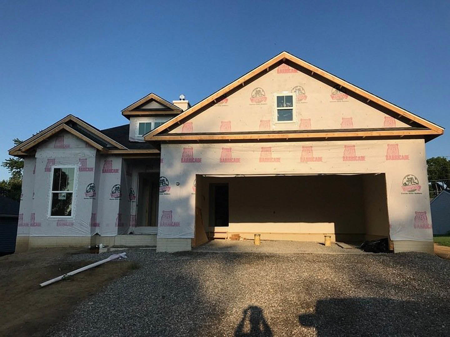 Partially built house with exposed framing, attached garage, white-framed windows, construction sign in window, white pipe on dirt ground, person’s shadow, blue sky overhead