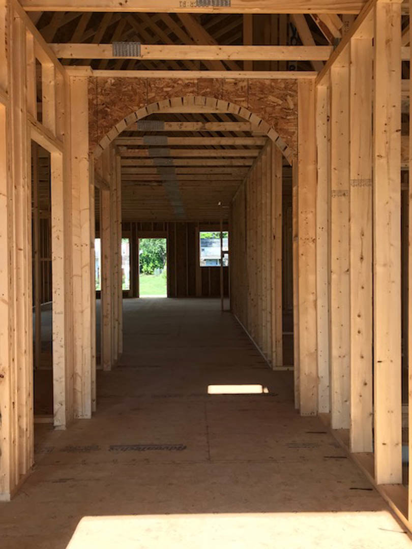 Hallway under construction featuring exposed wooden beams, arched doorways, and a wood arch; light shines on the unfinished floor, with windows offering views of trees and water.