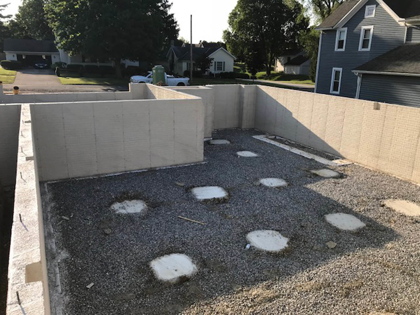 Concrete foundation with circular cutouts on gravel, surrounded by trees and sky, adjacent to a street curb and garage.