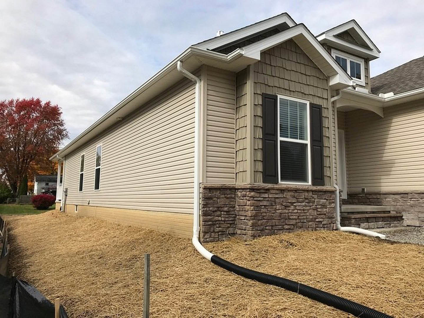 Two-story home with gray siding, stone accent wall, large windows with white blinds, red-leafed tree and bush, garden hose on front lawn, covered porch, shingled roof.
