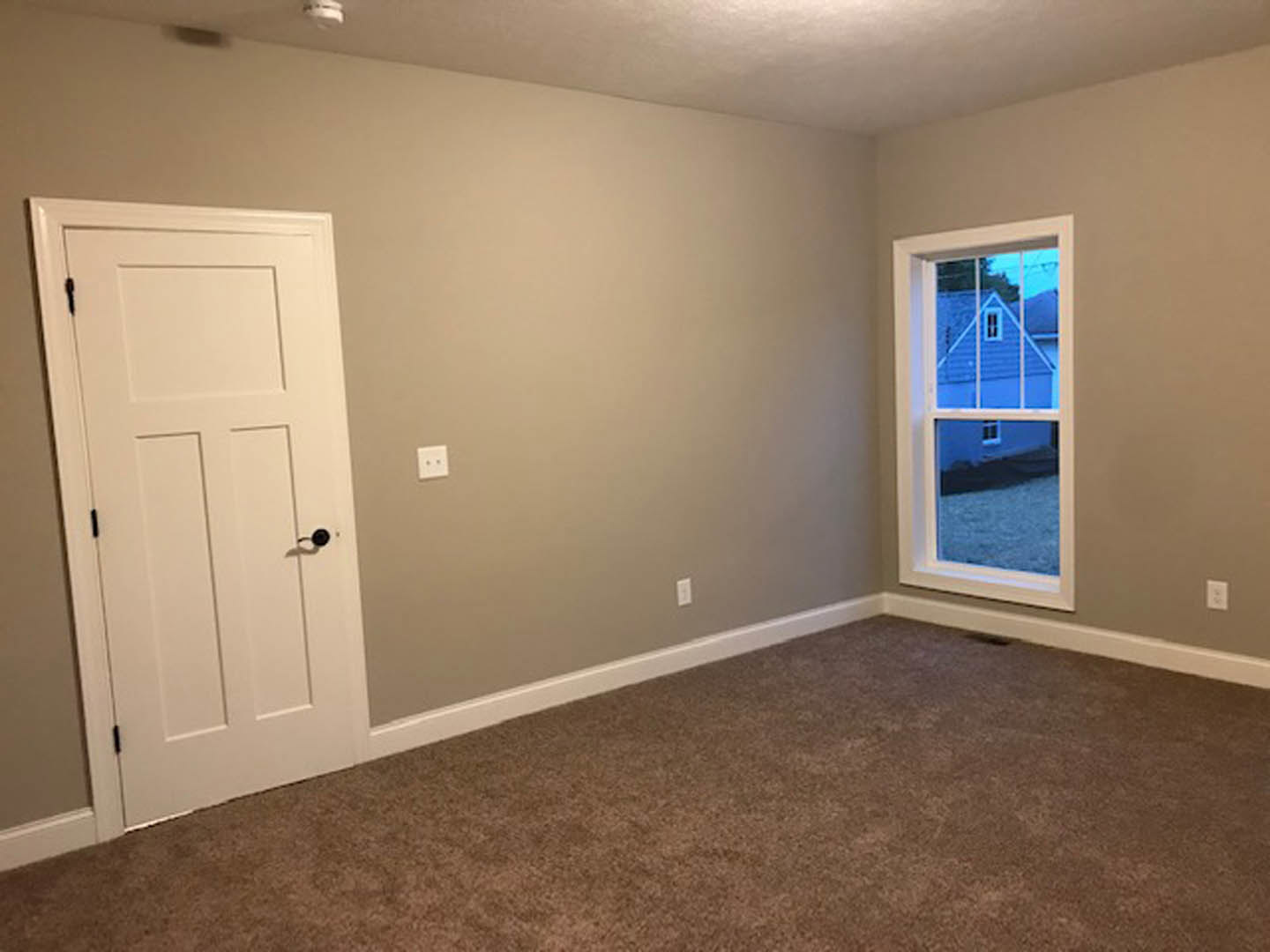 Brown carpeted room with white plaster walls, white door featuring a black handle, large window overlooking neighboring house, and white wall outlet.