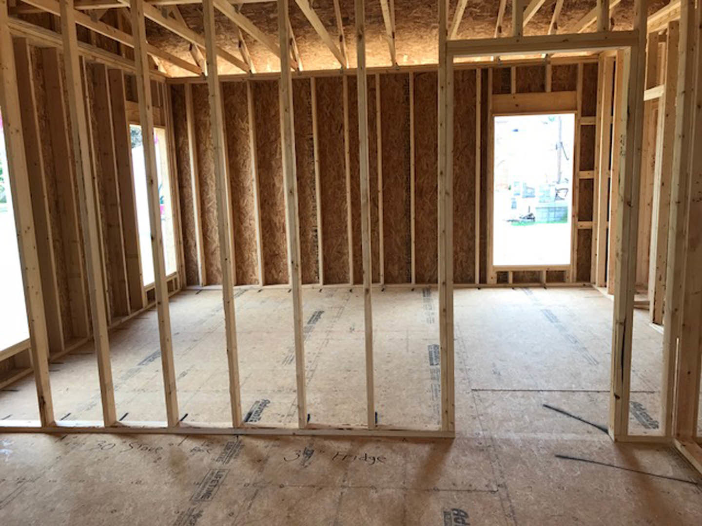 Wood-framed room under construction with exposed beams, unfinished floor, and large window offering view of neighboring building