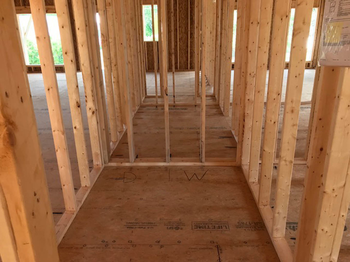 Living room with exposed wood ceiling beams, large window, unfinished wooden floor with handwritten markings, and construction materials scattered on the surface.