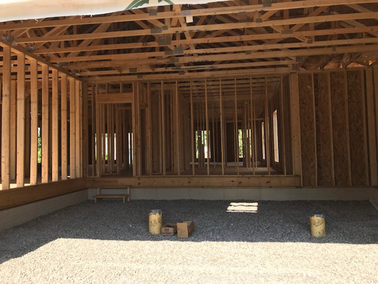 Wood-framed house under construction with exposed beams, gravel floor, and scattered boxes on the ground