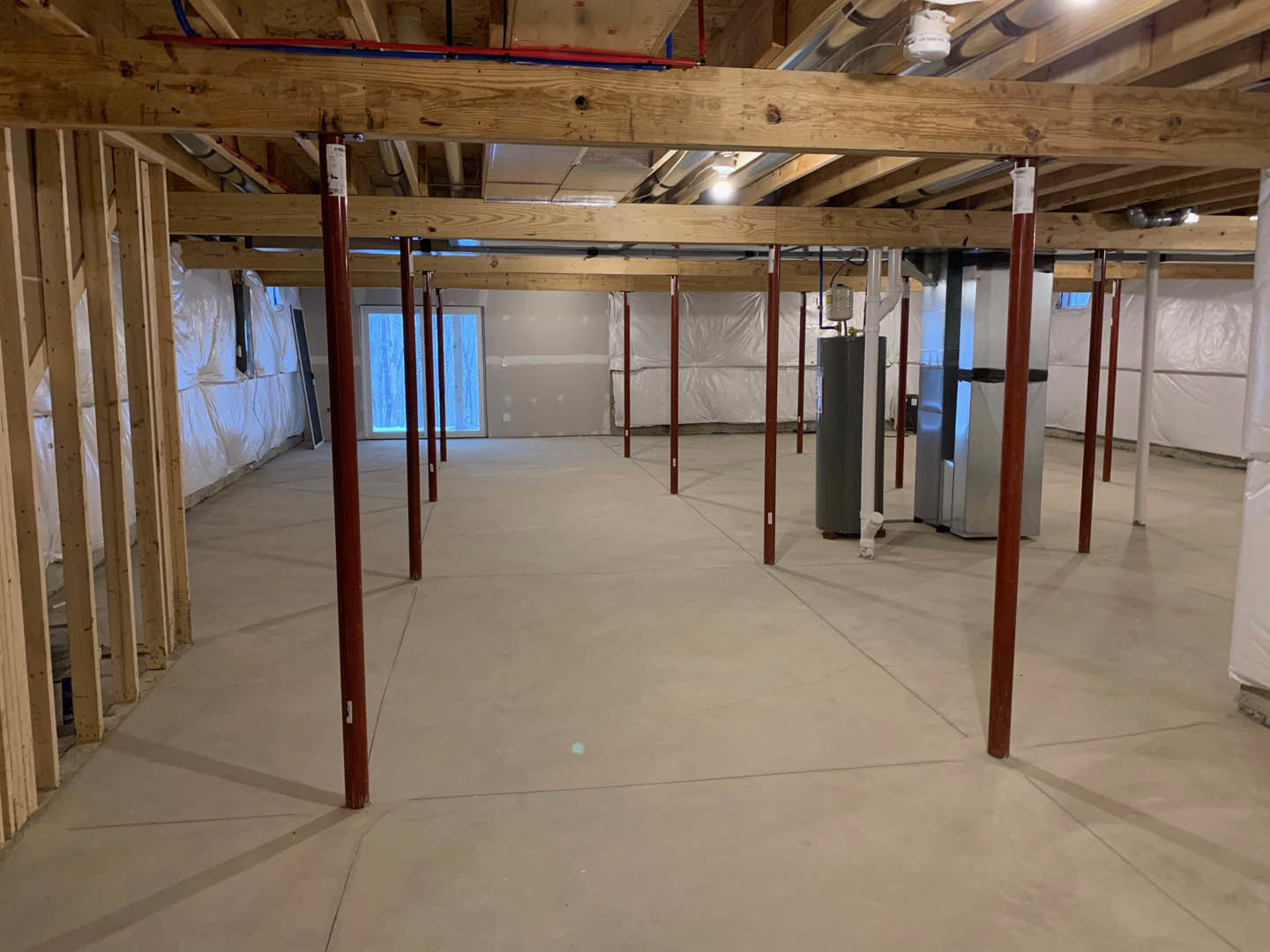 Exposed wooden ceiling beams and metal pipes above a concrete floor, with a window framed by vertical wooden poles and a grey structural pillar in the foreground