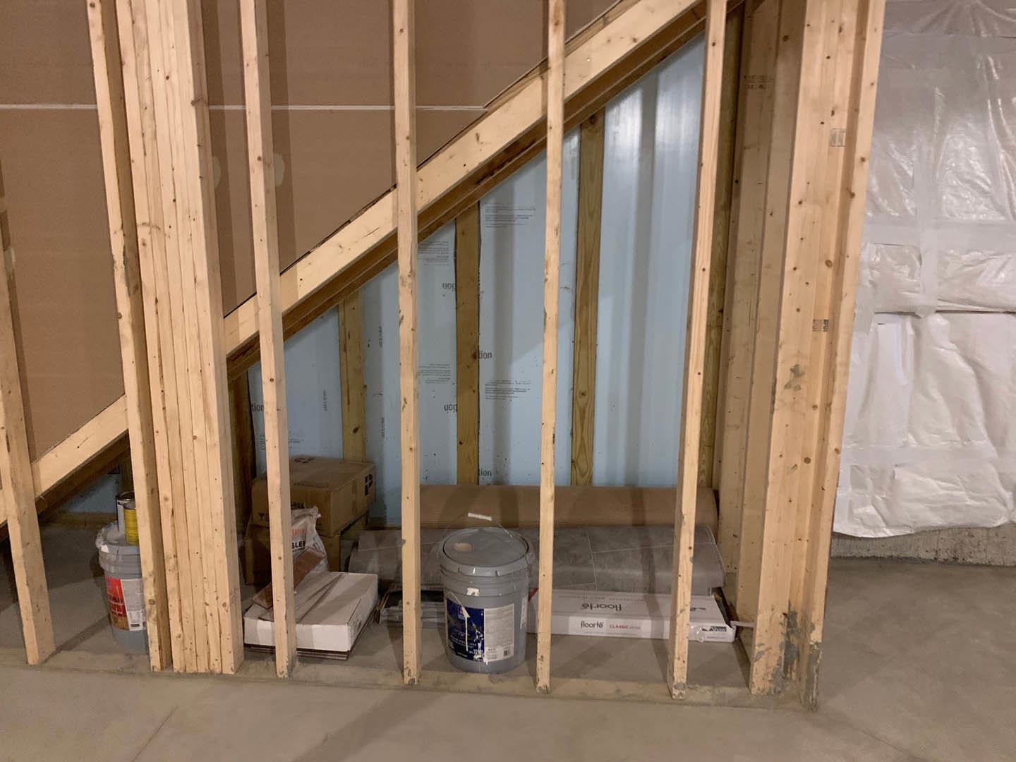 Unfinished wooden staircase frame with exposed lumber, paint bucket and white plastic bag nearby, white box resting on wooden surface, building insulation visible on walls