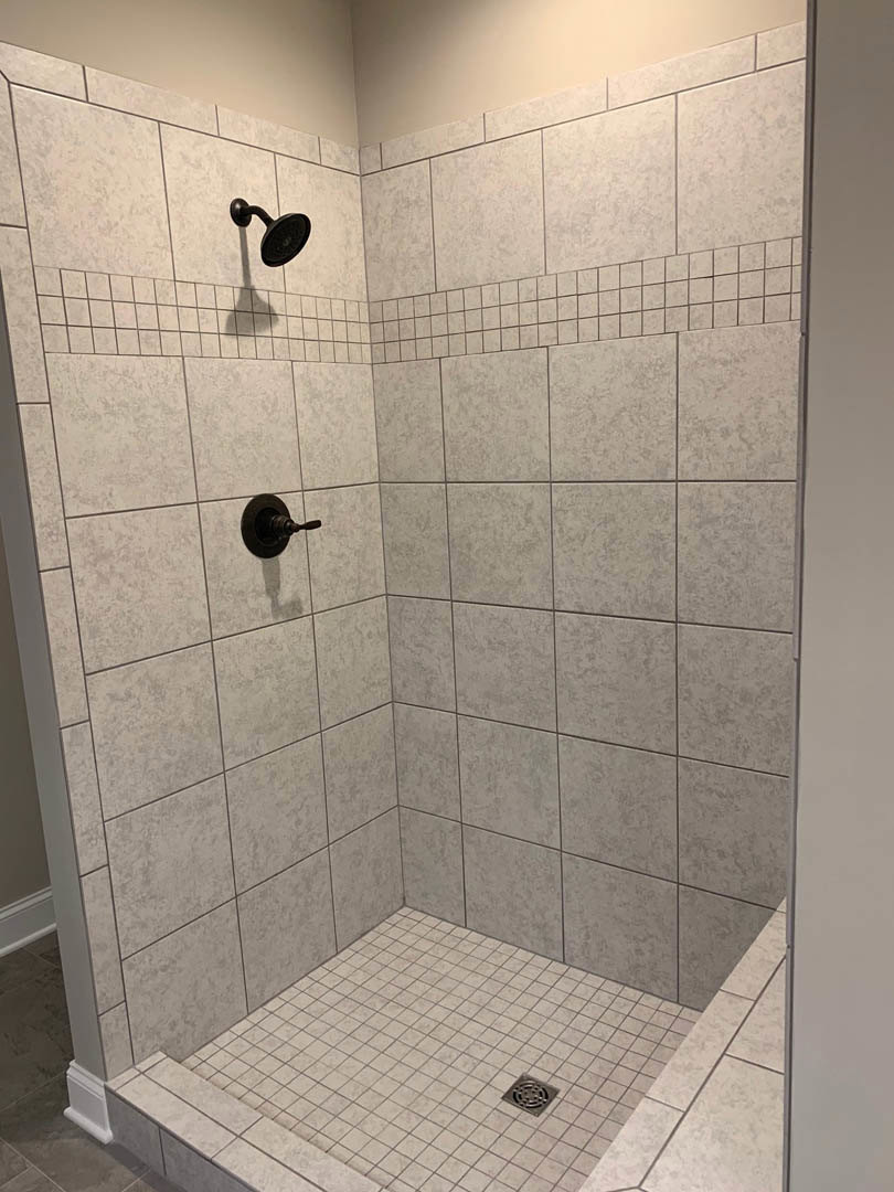 Modern bathroom shower with black shower head, white tile walls, tile floor with central drain, and nearby wall switch