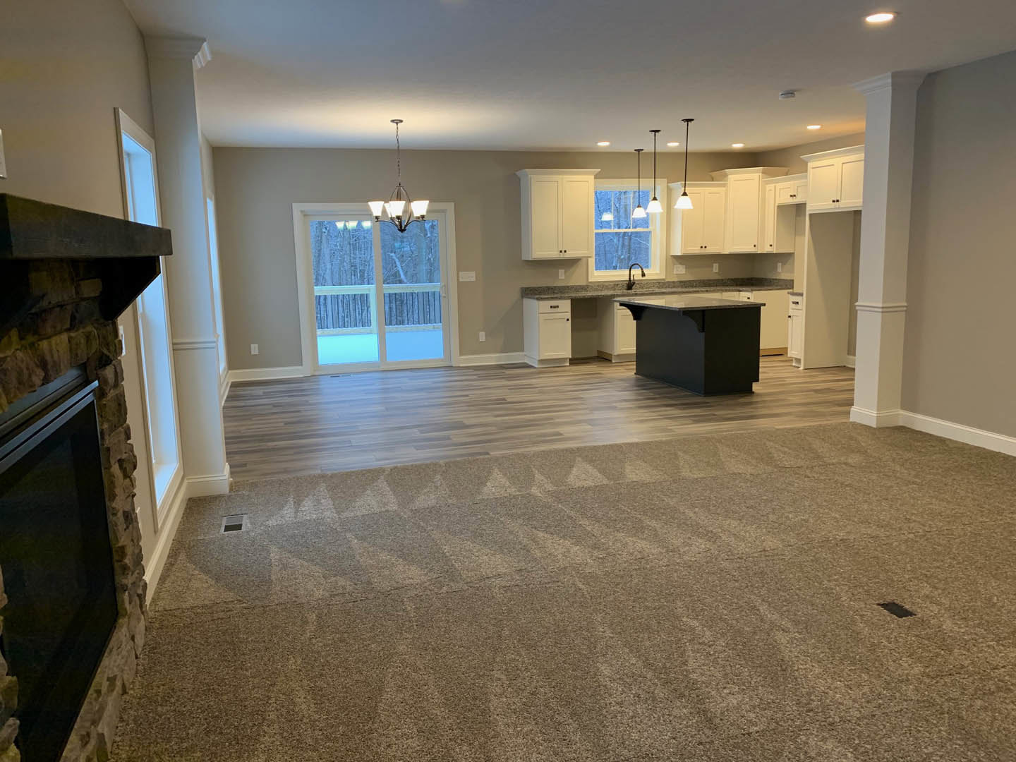 Open living room with stone fireplace, light tile flooring, modern chandelier, and adjacent kitchen featuring marble countertops and white cabinetry