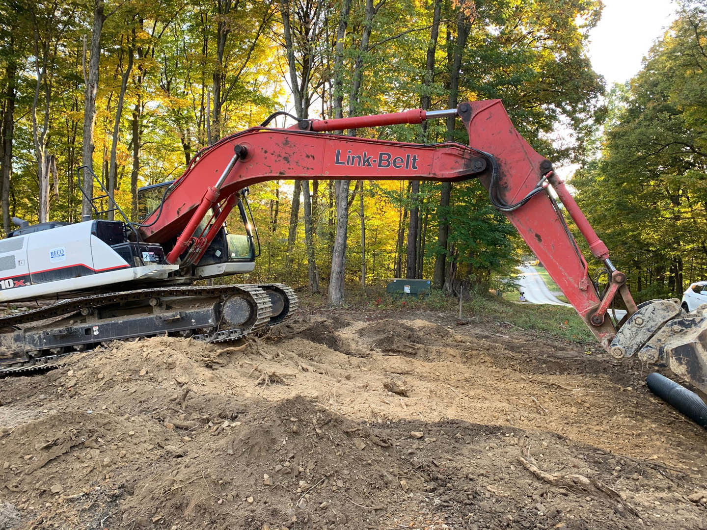 Red excavator with extended arm digging into soil, surrounded by dirt piles and construction equipment, trees visible in background