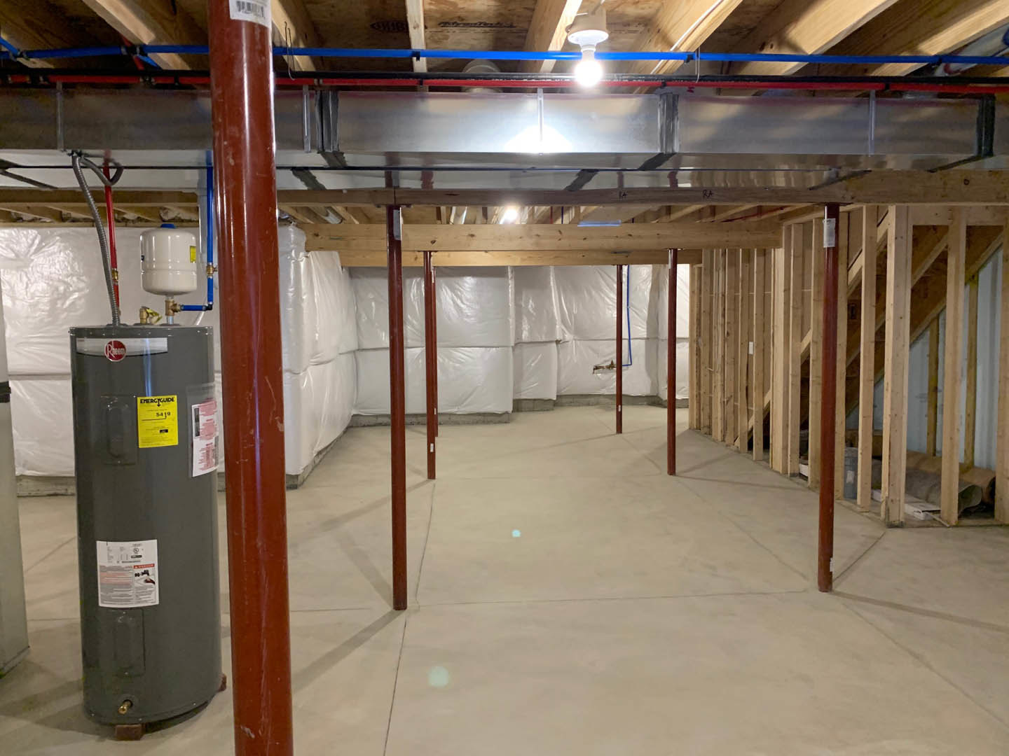 Utility room with exposed metal pipes along ceiling and walls, black cylinder tank, yellow caution sign on metal surface, white container with blue lid, and white and pink paper