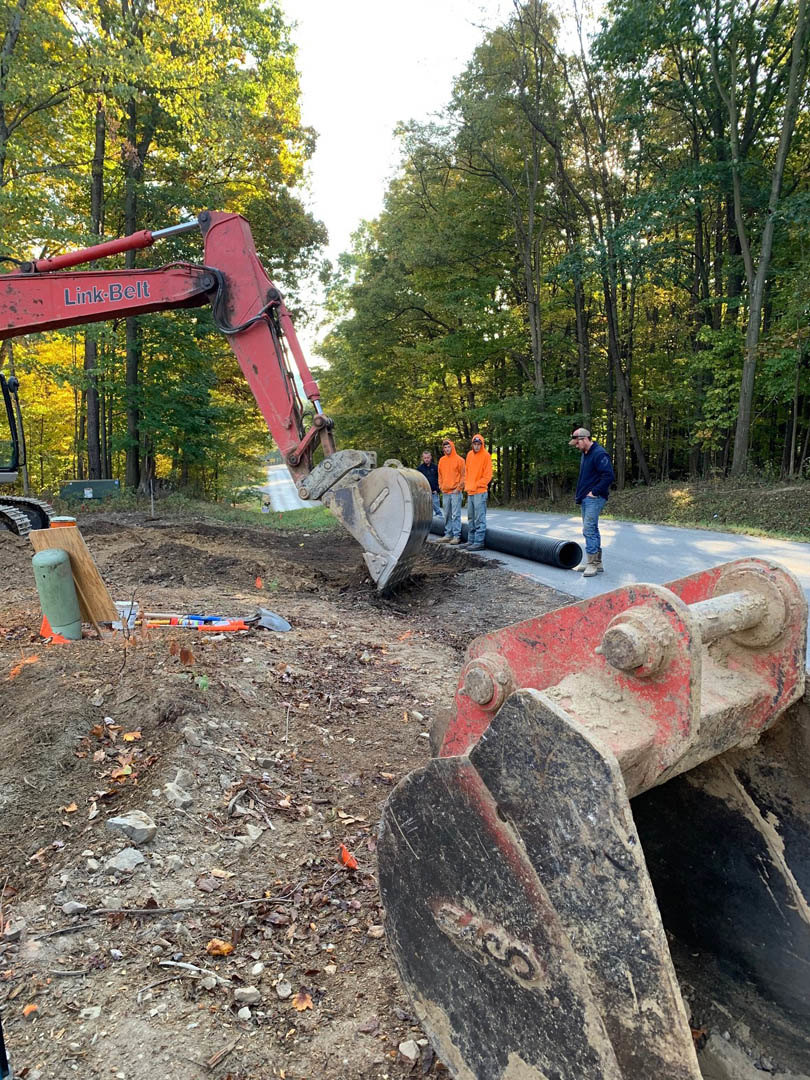 Group of construction workers in orange hoodies standing on soil near a red excavator arm, green striped pole, and bucket, with trees and blue sky in the background