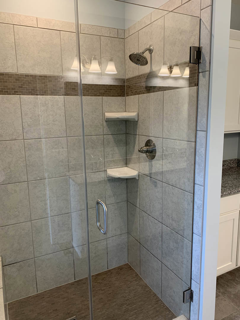 Glass shower enclosure with chrome hardware, white tile flooring, wall-mounted shower head, and partial views of a brick accent wall and modern light fixture.