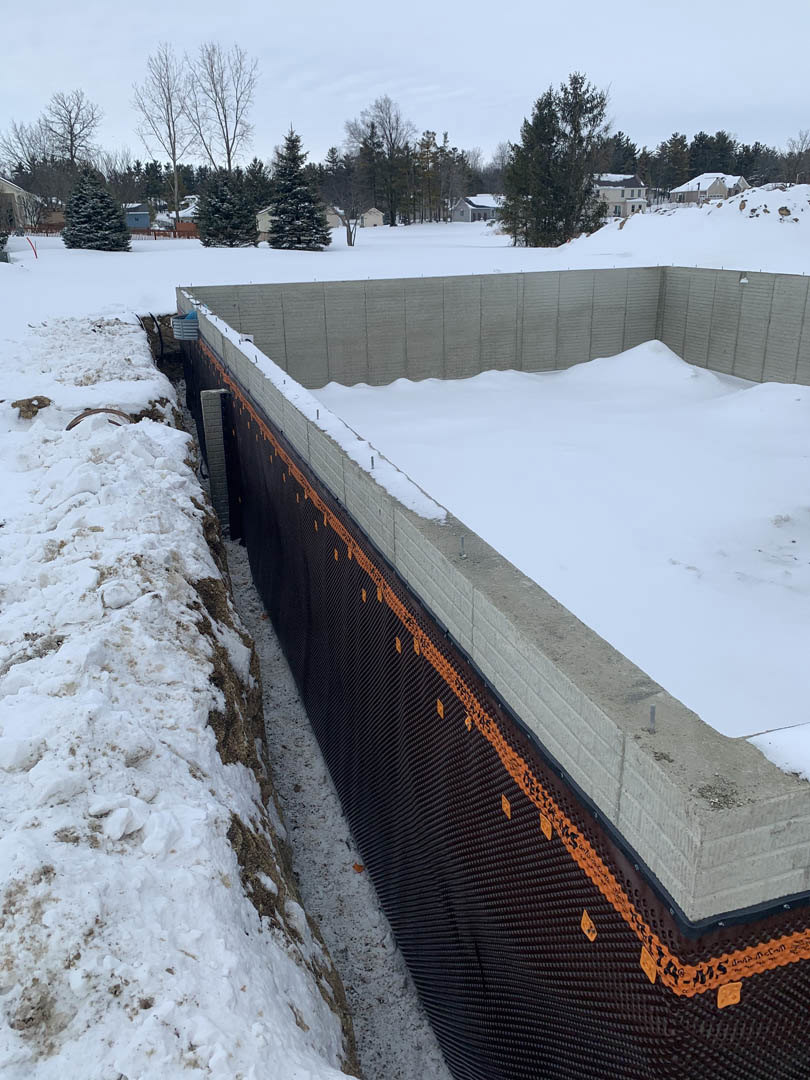 Concrete foundation under construction with foam insulation, surrounded by snow-covered ground and trees, winter sky overhead
