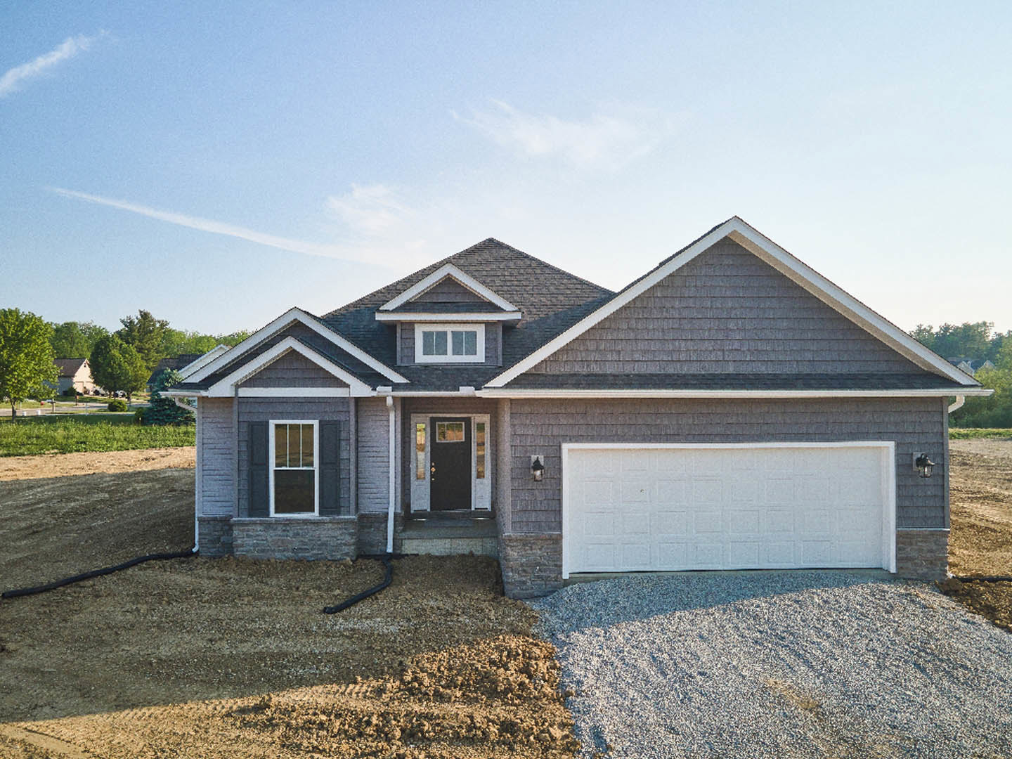 Two-story home with light siding, black front door with glass panel, attached garage with white door, gravel driveway, barred window, and surrounding trees under partly cloudy sky