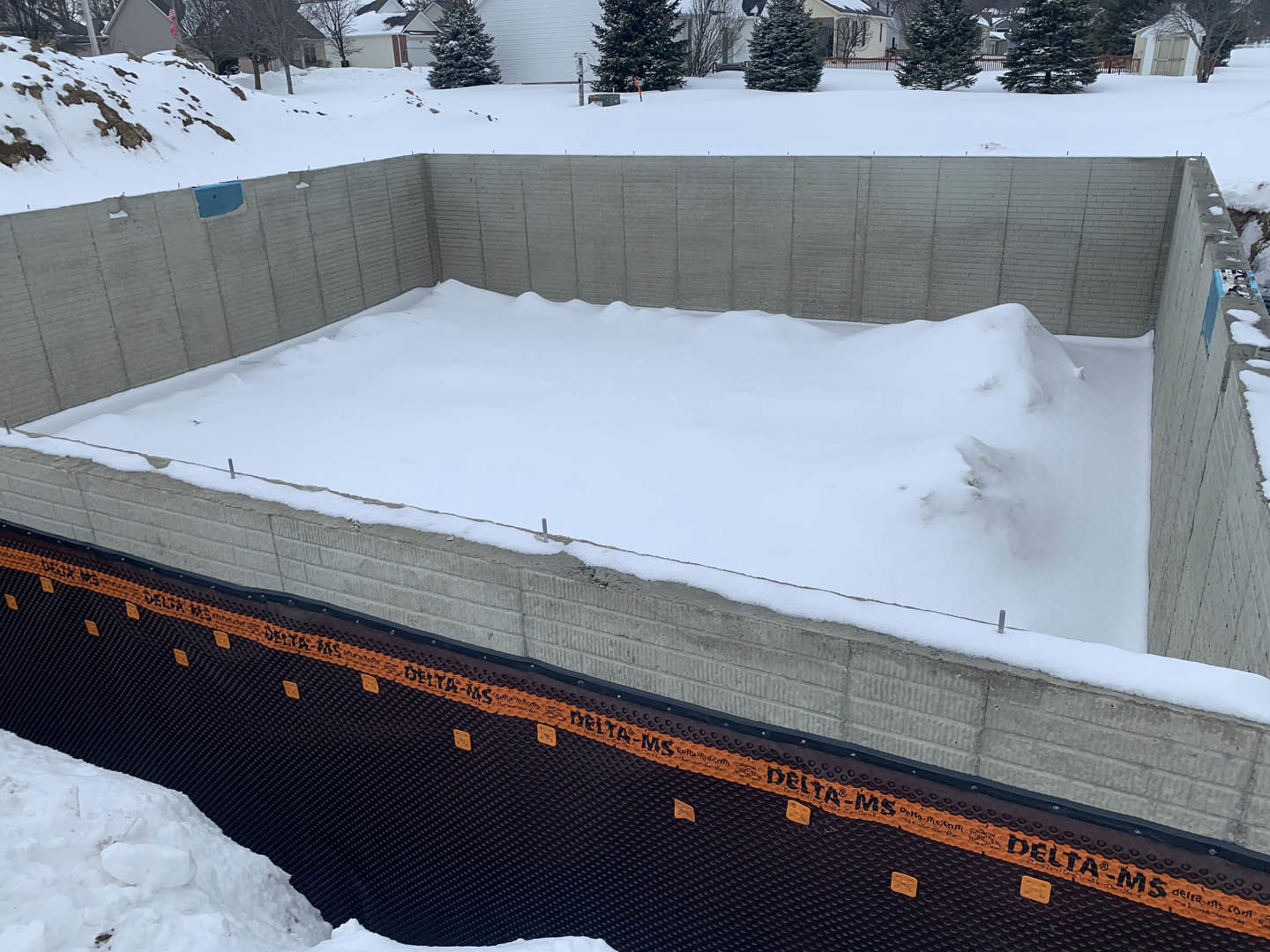Snow-covered concrete foundation with black and orange tape on the wall, blue rectangular object with central hole, tree and snowy ground in a winter landscape
