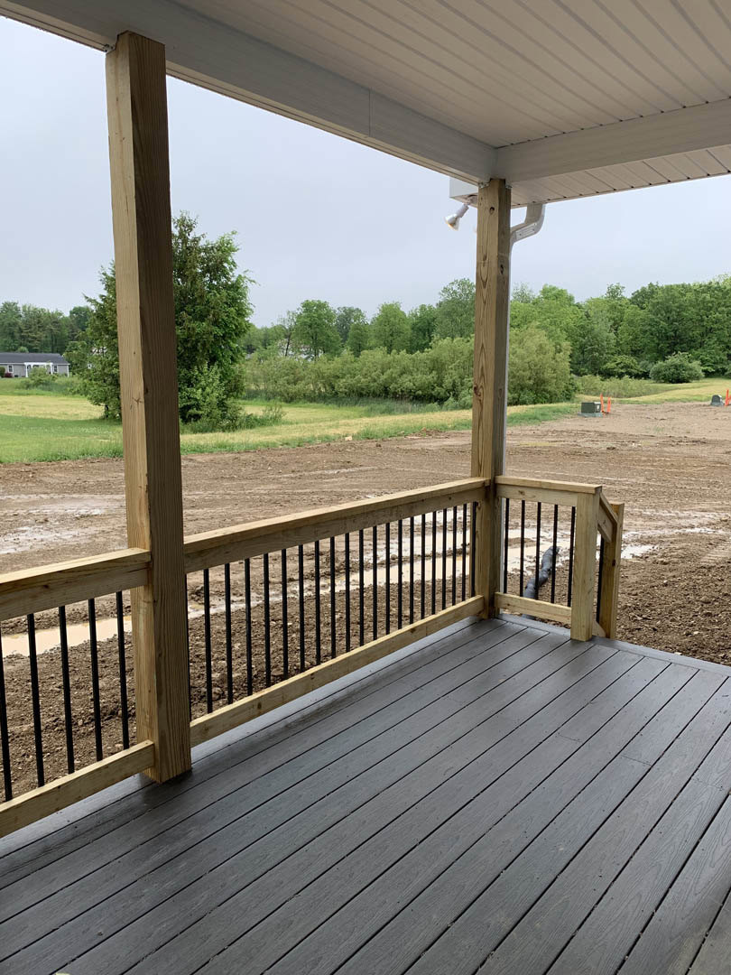 Wooden deck with horizontal fence overlooking a dirt field, tree with green leaves nearby, partial view of house exterior and window.