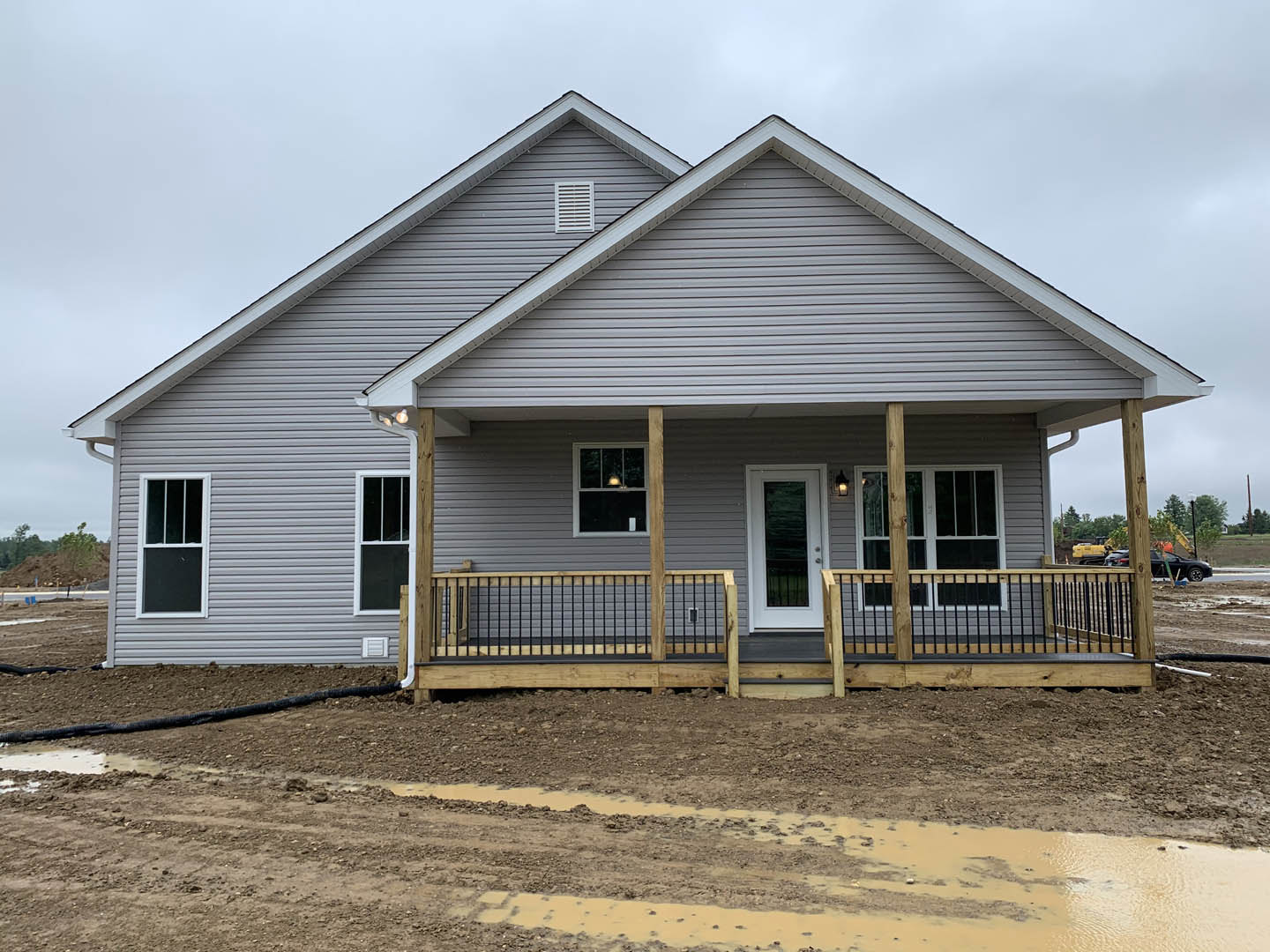 Partially built house with exposed wood framing, white siding, porch area, white vent on exterior wall, door with white trim, metal fence supported by wood posts, dirt path leading