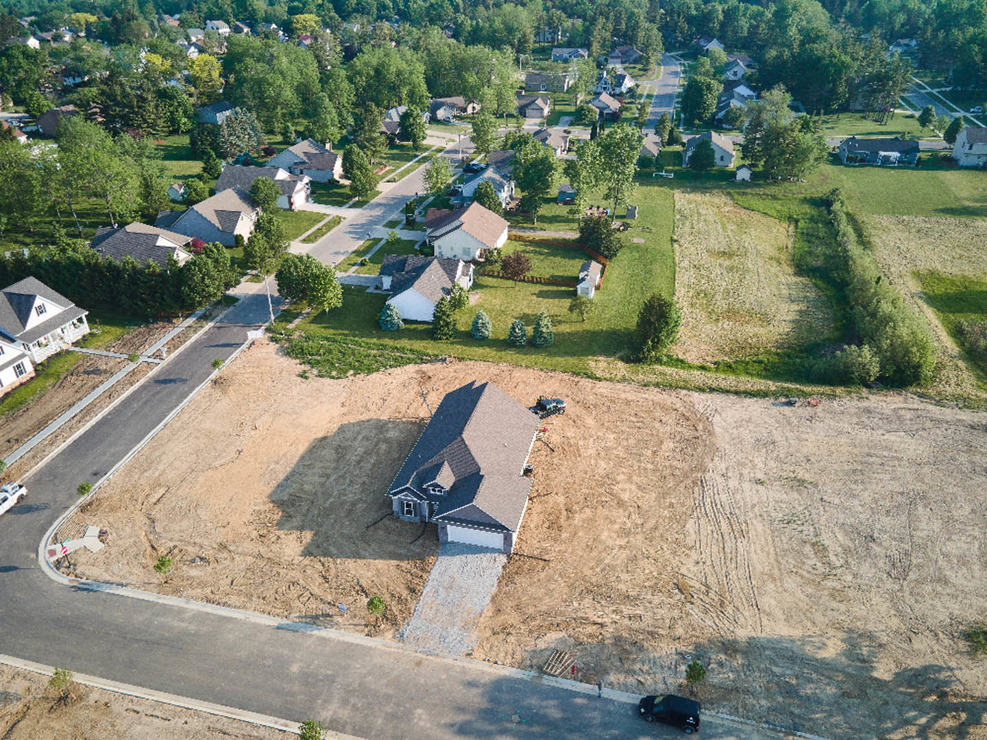 Aerial view of suburban neighborhood with multiple houses featuring garages and porches, tree-lined streets, grassy yards, a car driving on the road, and a crosswalk sign visible
