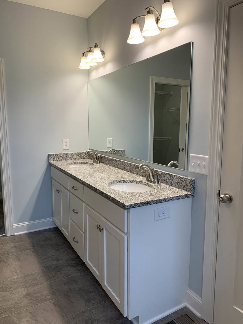 Bathroom with granite countertop, two undermount sinks, large framed mirror, white cabinetry, chrome faucets, and tiled floor