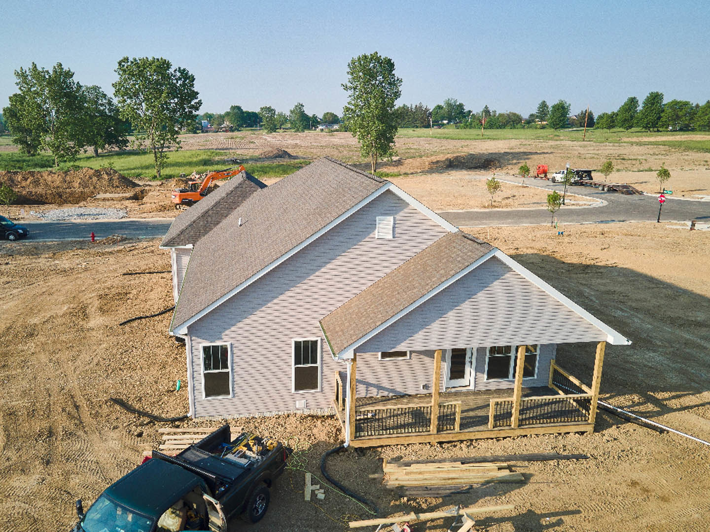 Partially built house with exposed framing, white and black window frames, construction truck parked in front, grassy yard, mature trees surrounding the site, temporary fencing
