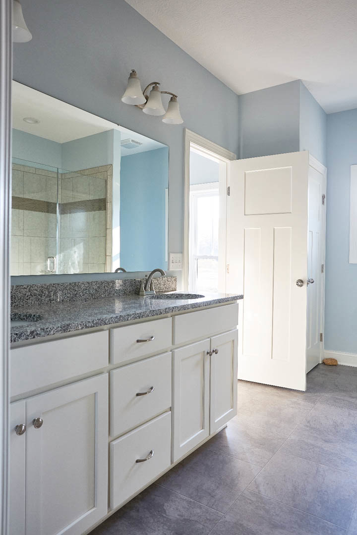 Bathroom with white vanity, rectangular mirror, three-bulb light fixture above, glass shower door, light gray tile flooring, and white door with sunlight streaming through.