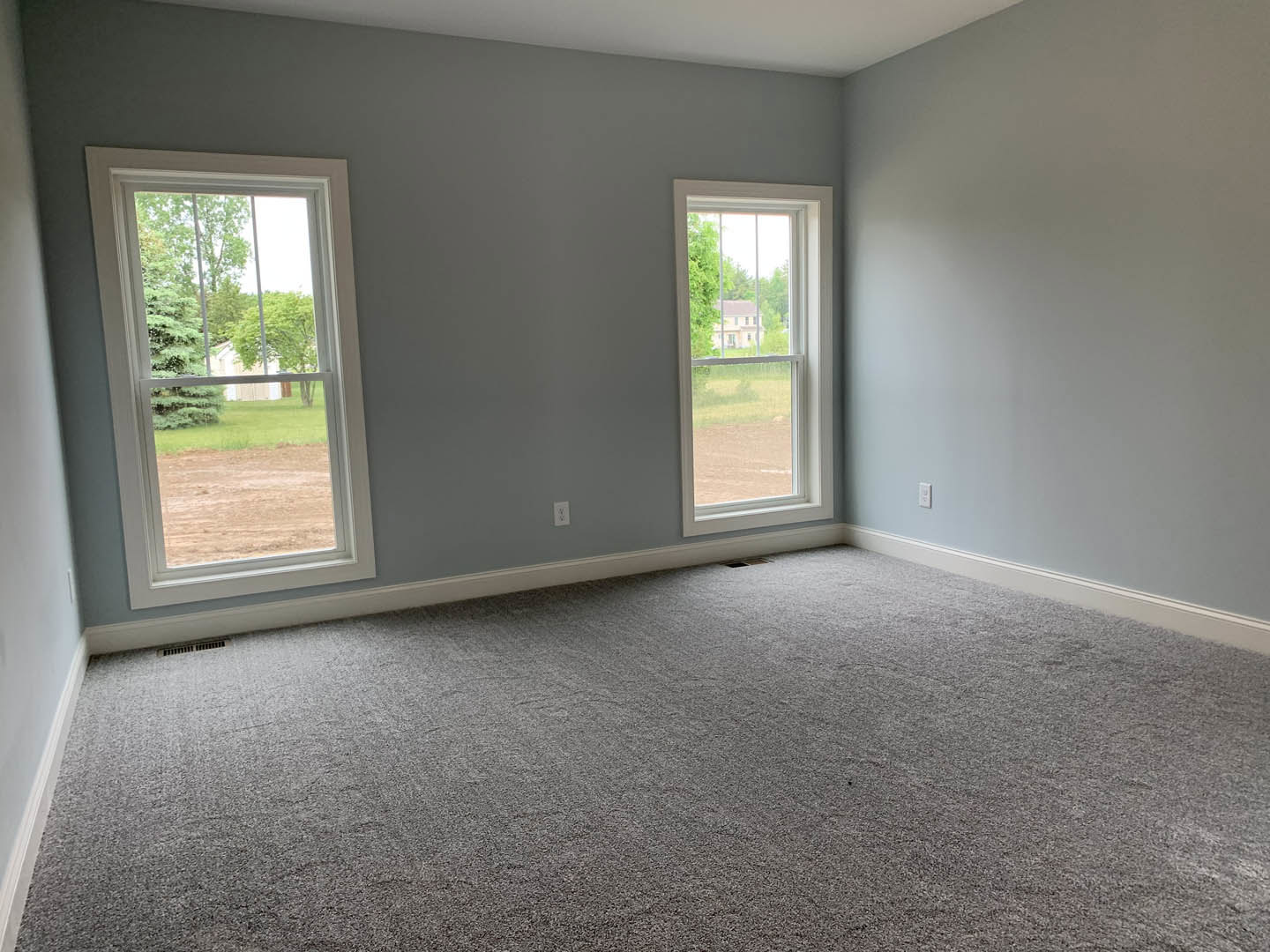 Neutral-toned carpeted room with two large windows, white walls, open door, and views of trees and a neighboring house outside