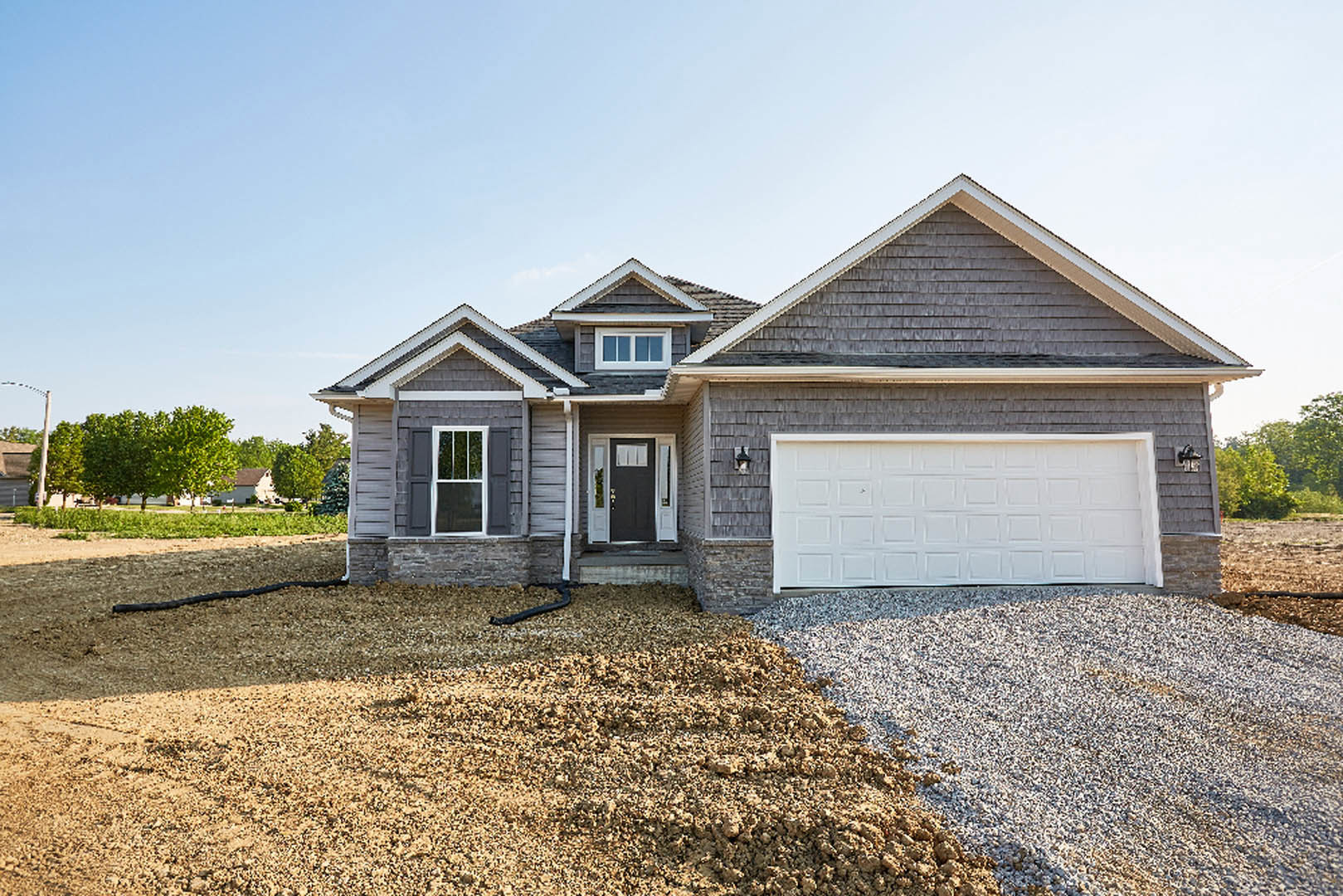 White garage door and black roof on a modern home, black entry door with window, white-framed windows, driveway with gravel and dirt pile, tree and sky in background