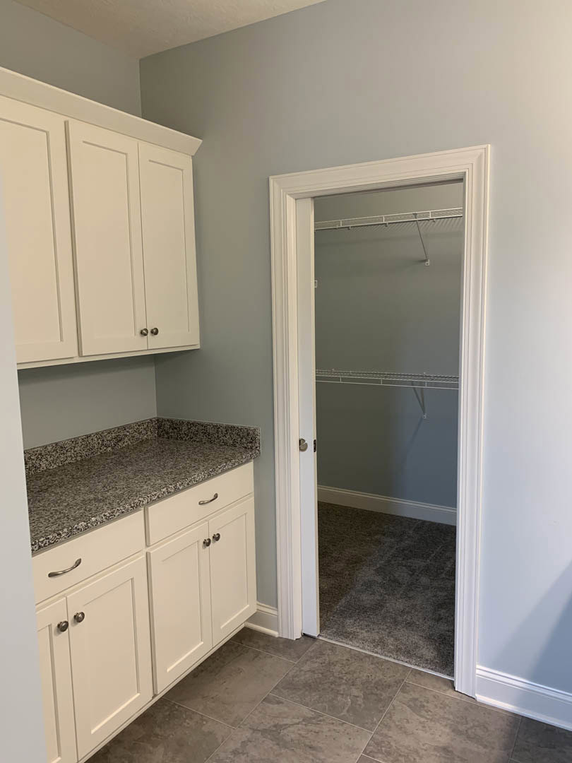 White door with black handle opens to a room featuring tile flooring, white cabinetry with drawers, a countertop, and a closet with a white shelf.