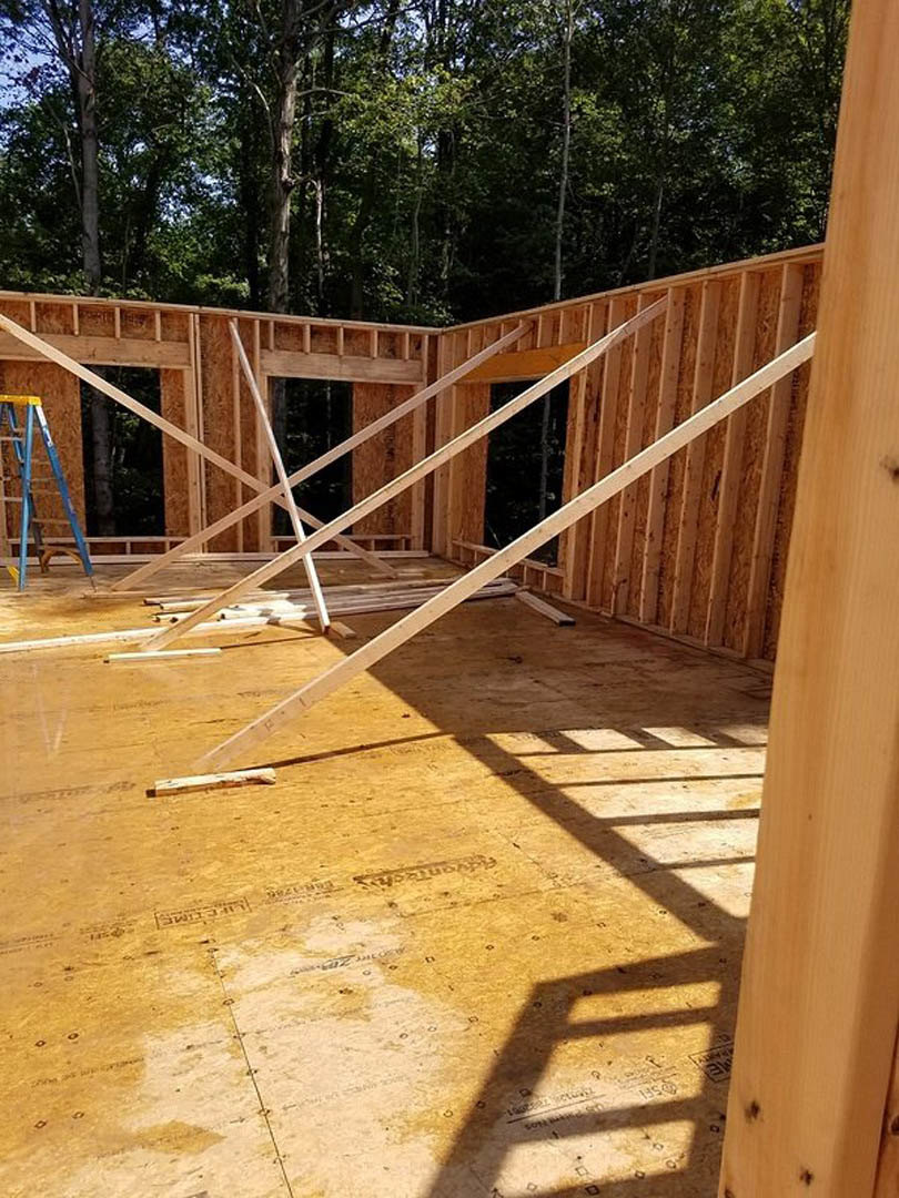 Wood framing and exposed beams of a house under construction, blue ladder leaning against partially built brick wall, surrounding trees visible in background