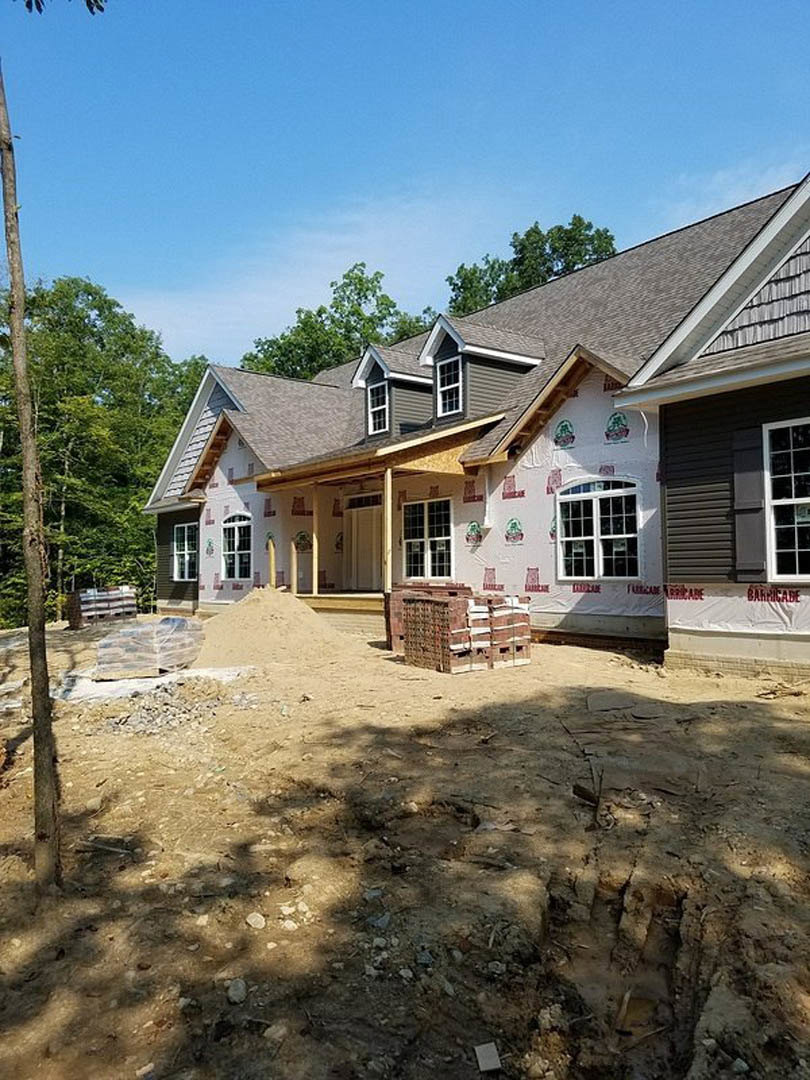 Two-story house under construction with exposed brick walls, pile of sand in front, scattered building materials, and surrounding trees.