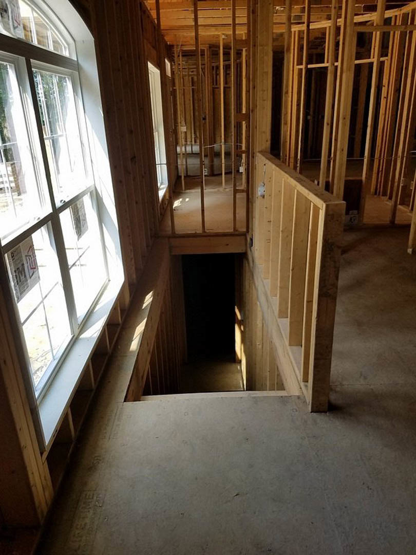 Unfinished room featuring exposed wooden beams, a window with papers taped to the glass, raw lumber planks, and a partially constructed staircase.