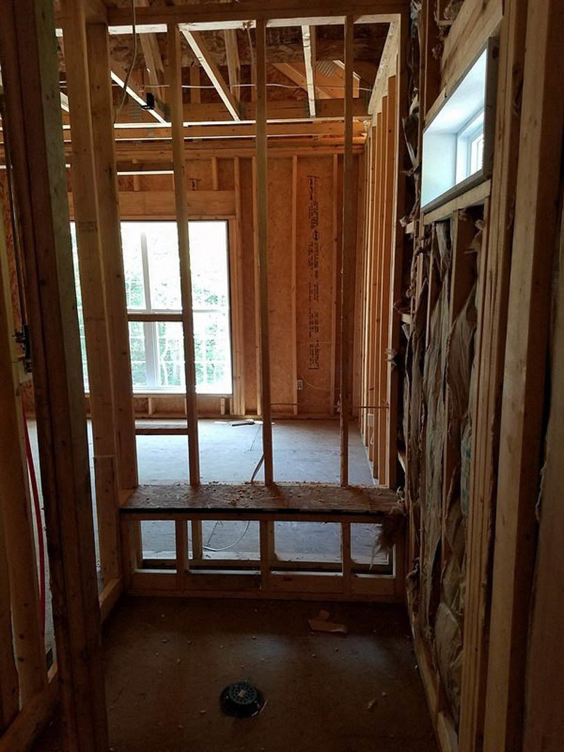 Sunlit room with white-framed window, wood beam ceiling, light-colored walls, and unfinished wood plank flooring with visible floor drain