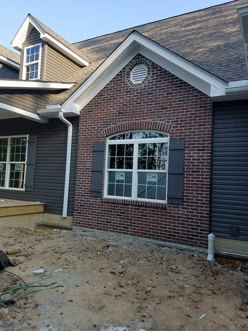 Red brick exterior wall with a multi-pane white framed window, dirt ground scattered with rocks, sticks, and a broken green wire