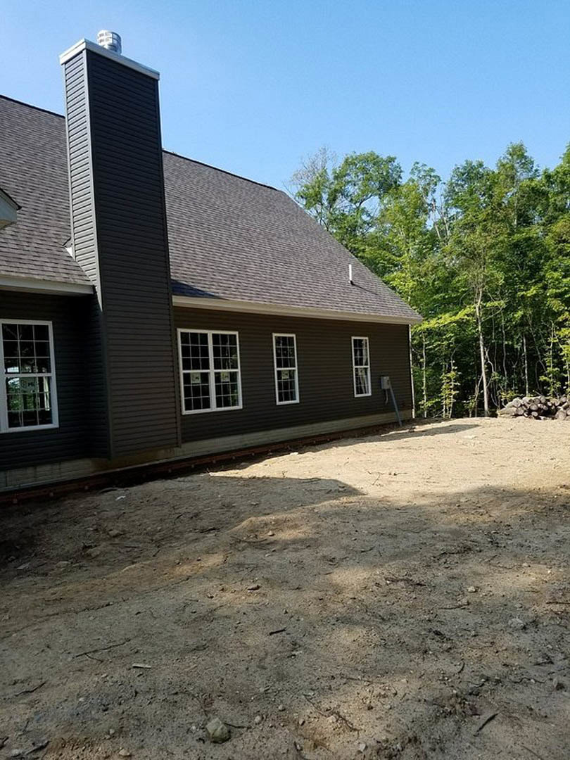 Two-story house with multi-pane windows, brick chimney, fenced dirt yard, and trees in the background