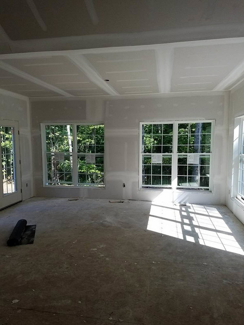 Bright room with multi-pane windows, white flooring, roll of black carpet, and door; sunlight casts shadows across the floor, trees visible outside.