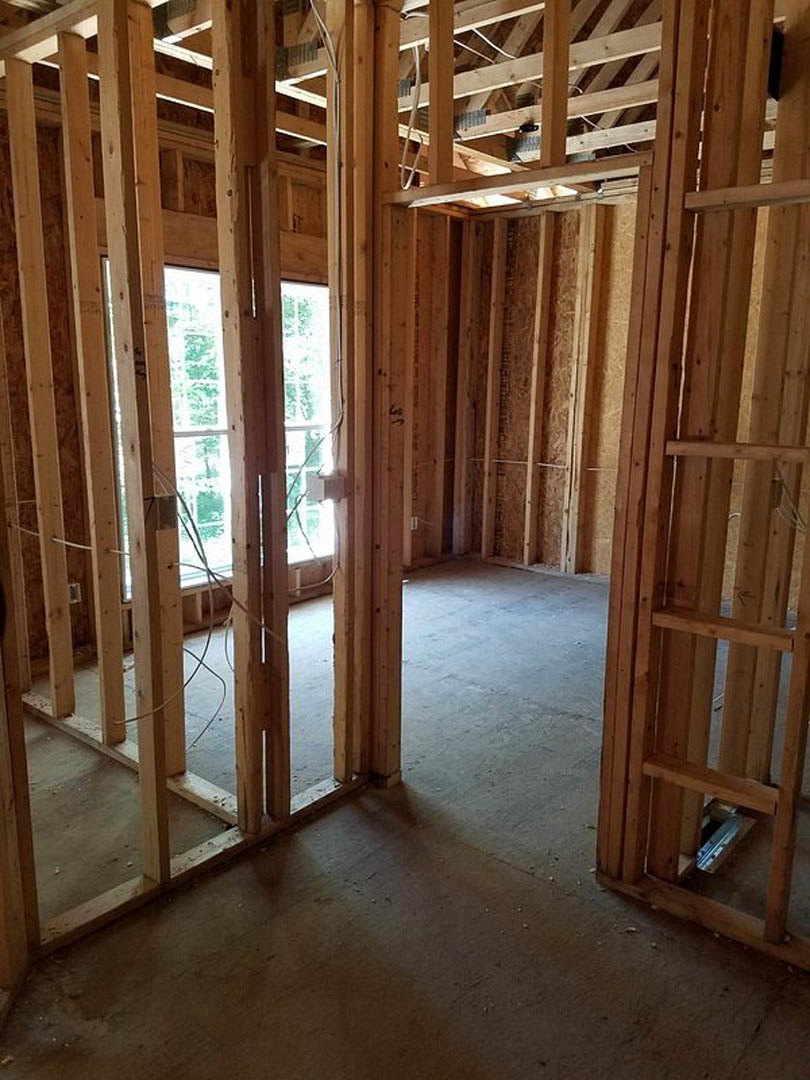 Unfinished room with exposed wood framing, visible beams, door opening, and multiple windows, some showing leafy green patterns outside; electrical wires run along the ceiling.