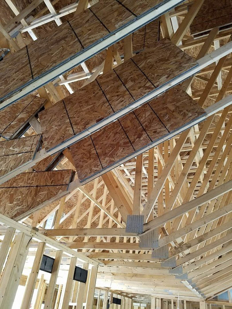 Exposed wood beams and boards forming a sloped roof structure inside a house, with a pile of plywood and construction materials visible.