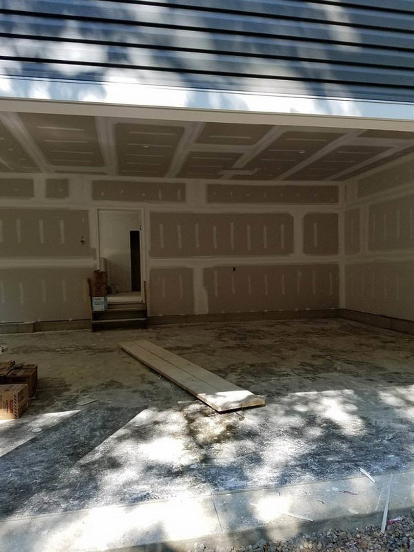 Wood plank resting on concrete garage floor beside white wall and black door, blurred window in background, close-up of cardboard box nearby.