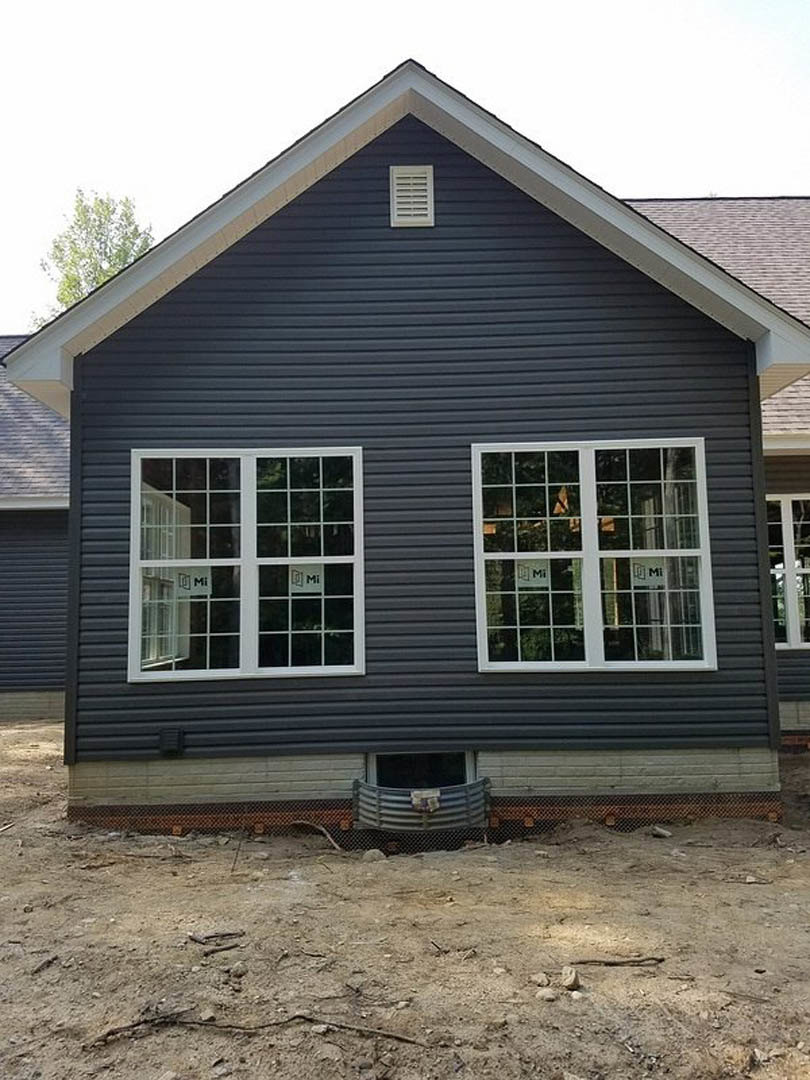 Partially built house with exposed wooden framing, several installed multi-pane windows, white exterior vent, and gray siding under a clear sky