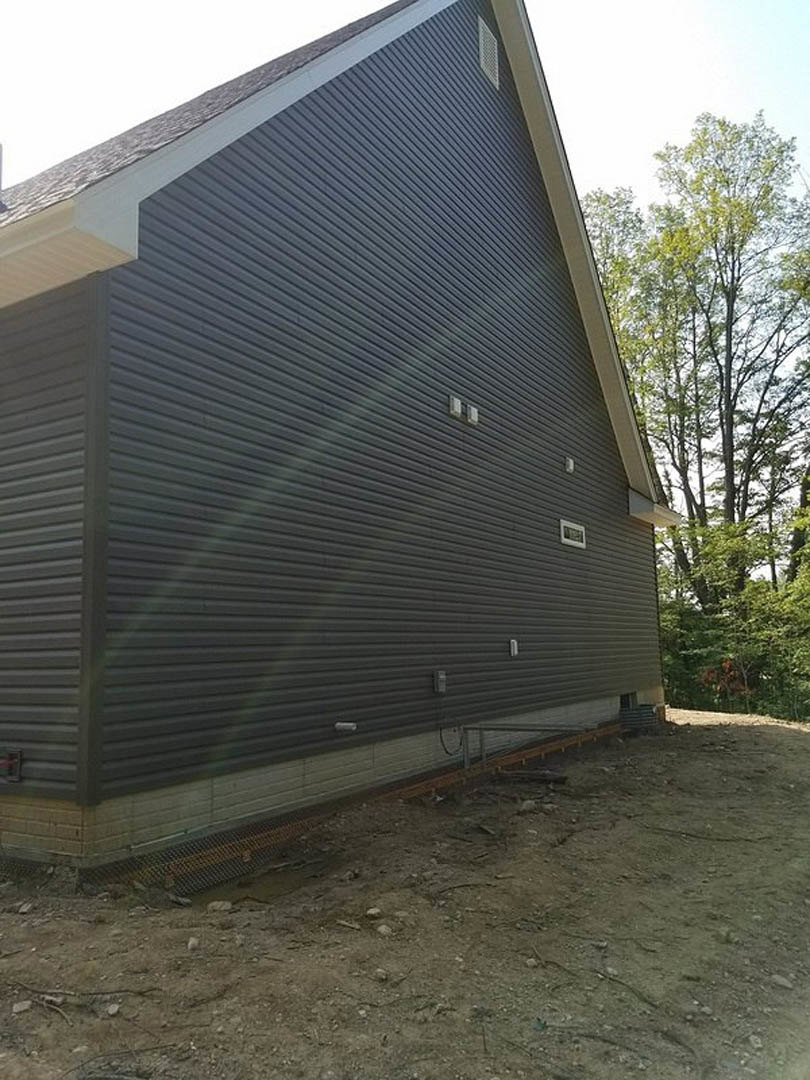 Modern custom home with light-colored siding, sloped roof, and large windows; side view shows dirt yard bordered by a metal fence and mature tree.