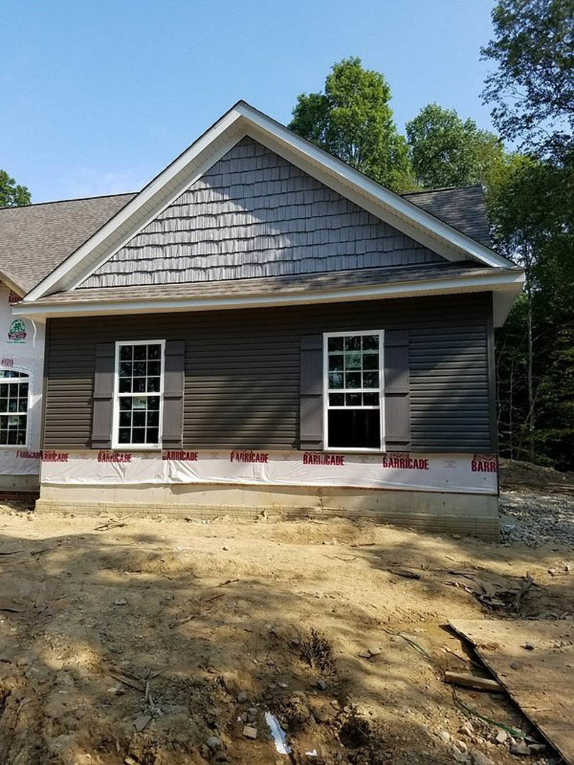 White metal roof atop a single-story home with light siding, fenced yard, dirt ground, large window displaying a sign, and mature trees in the background.