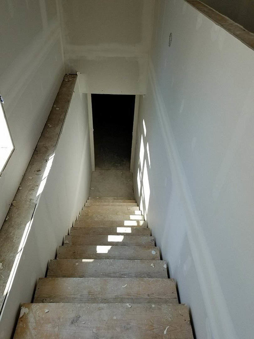 Wooden staircase with sunlight streaming across steps, black door with white frame at landing, grey plaster wall, composite handrail, light highlighting natural wood grain.