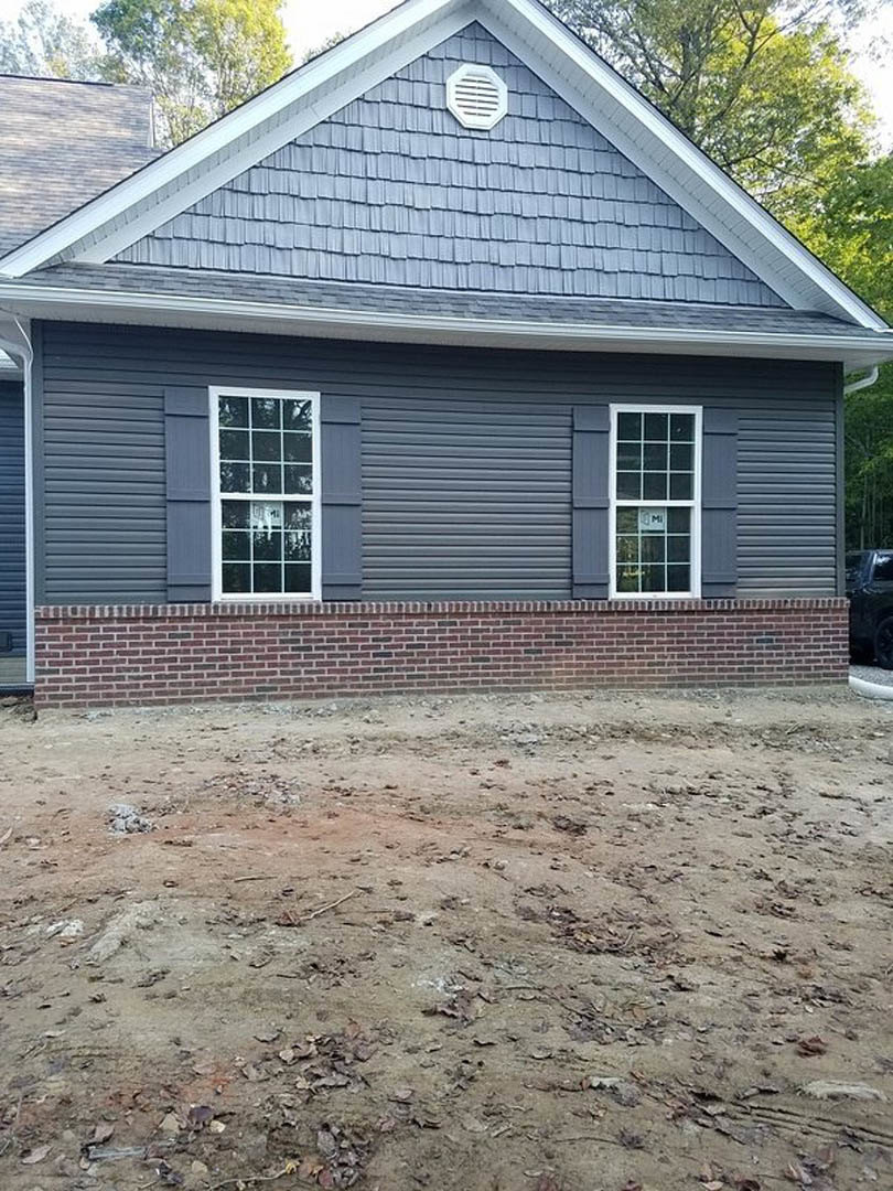 Brick exterior home with multi-pane window, black trim, wall vent, and dirt yard scattered with leaves.