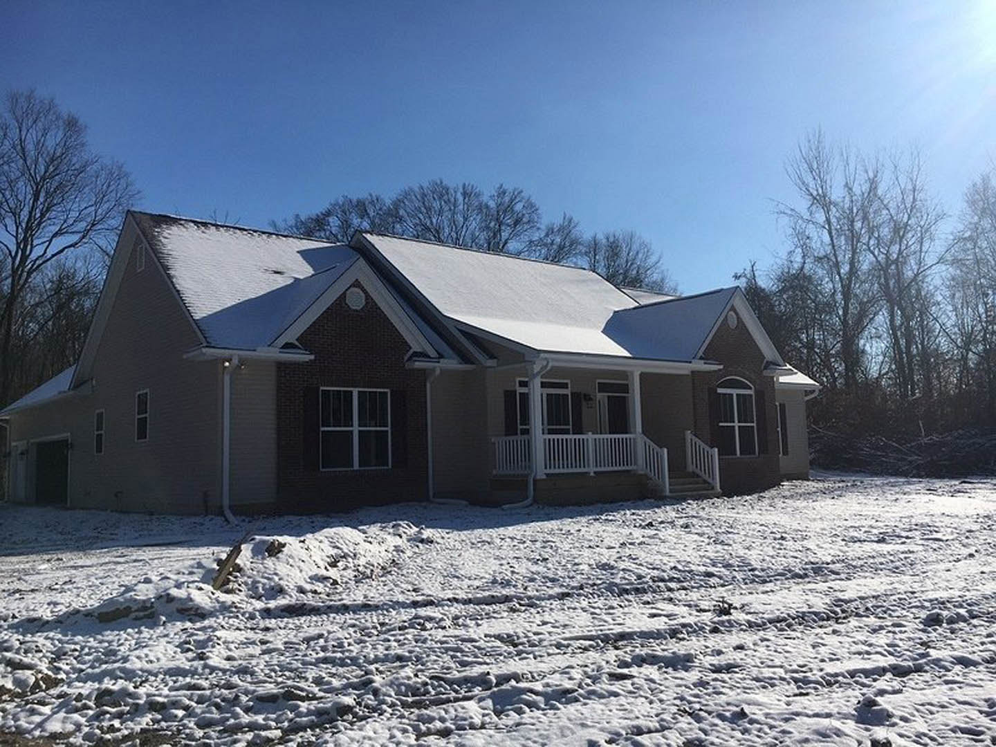 Two-story house with snow-covered roof and ground, white-framed windows, white porch fence, bare trees, and overcast winter sky