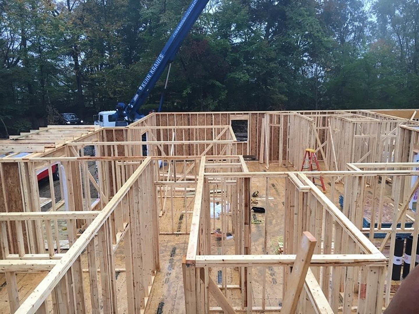 Wooden house frame under construction with crane lifting lumber, surrounded by trees and construction vehicles.