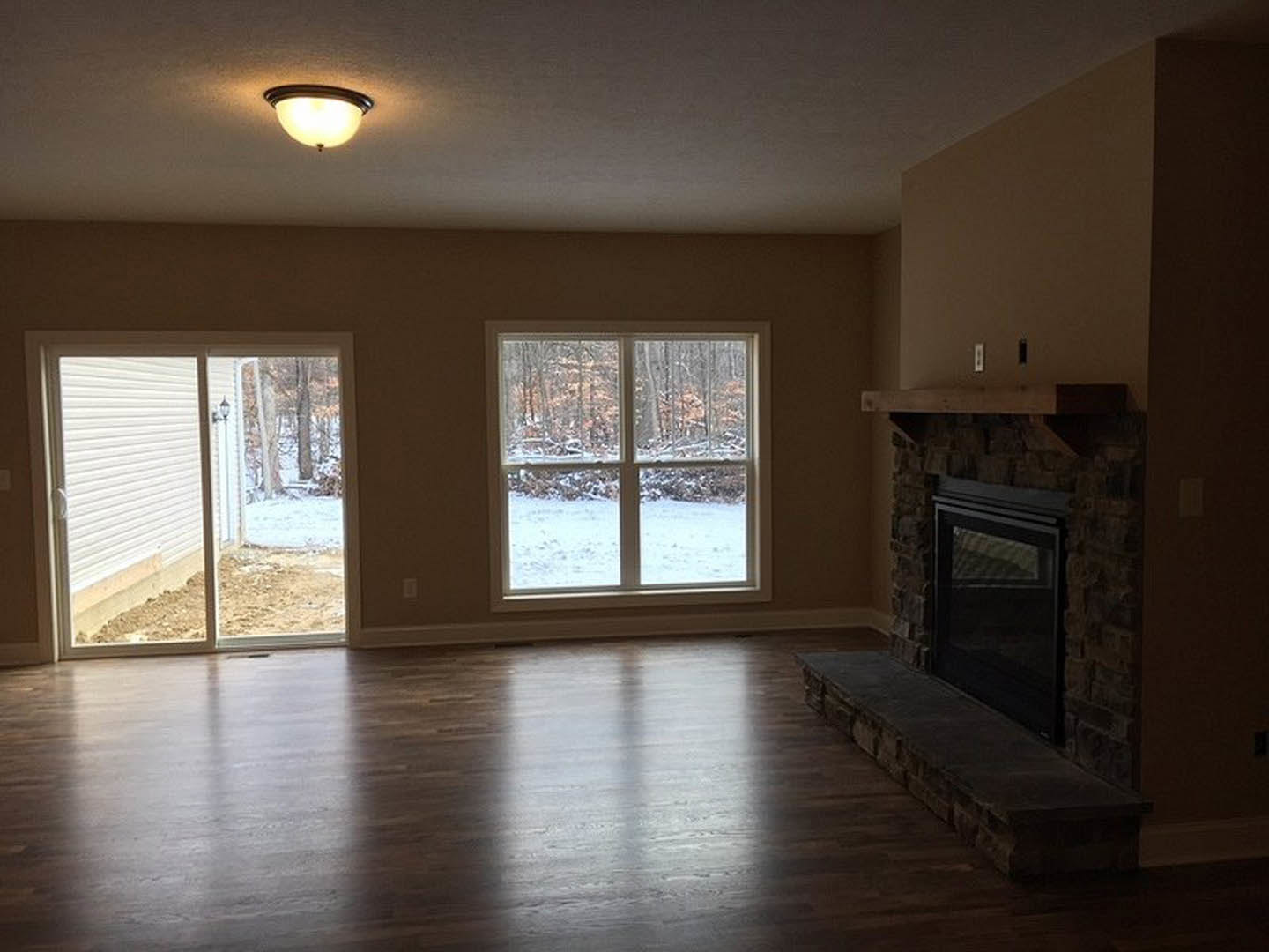 Living room with wood flooring, stone fireplace and ledge, large windows and sliding glass door overlooking snowy yard, ceiling light fixture visible