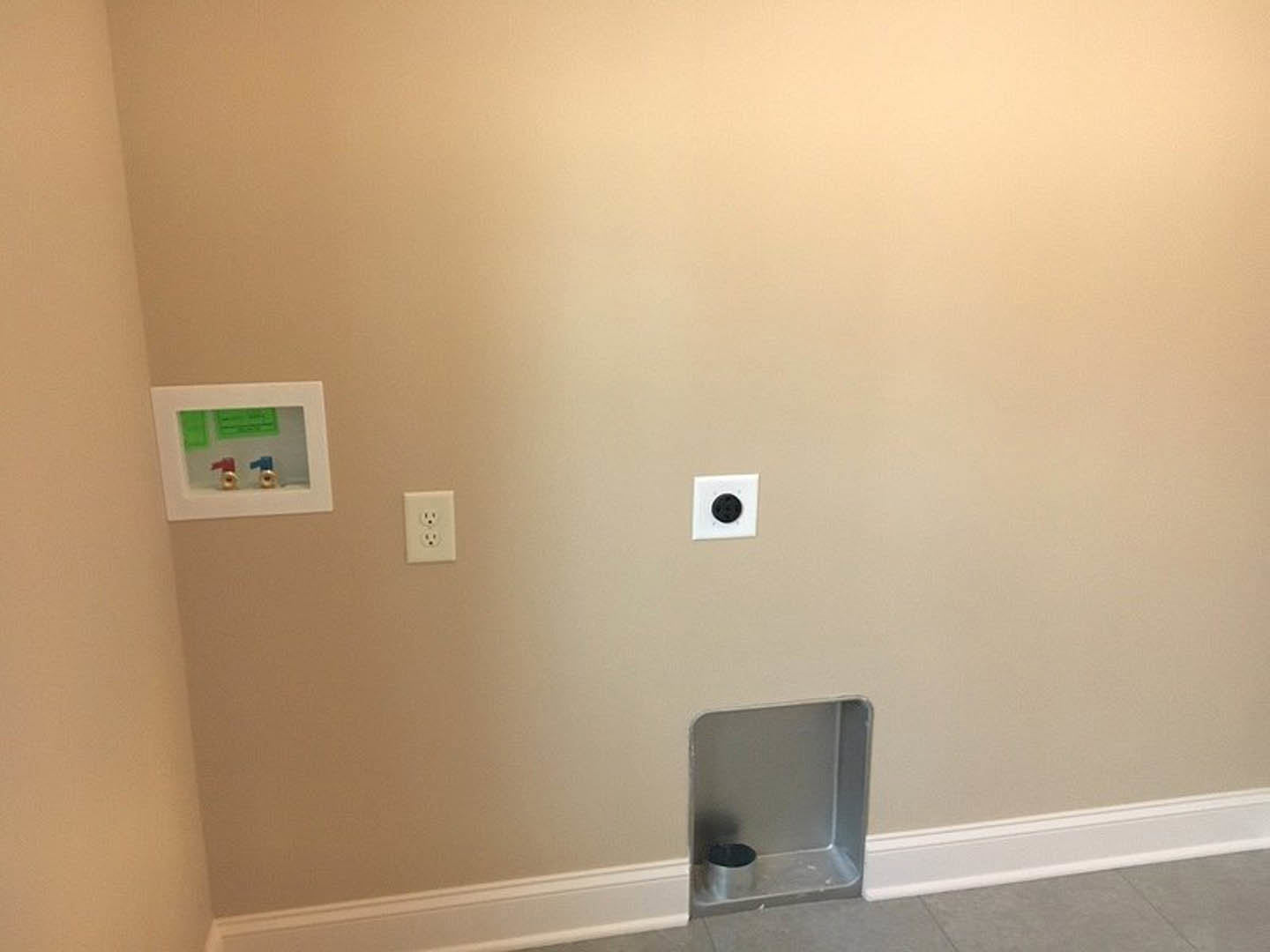 White plaster bathroom wall with a circular hole, exposed metal plumbing fixtures, blue and red faucet handles, electrical outlet, and black circle detail near the floor.